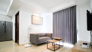 A modern living room with a sofa, a standing fan, a small table, and a TV on a stand. - Exclusive And Spacey 3Br At Grand Sungkono Lagoon Apartment (Surabaya)