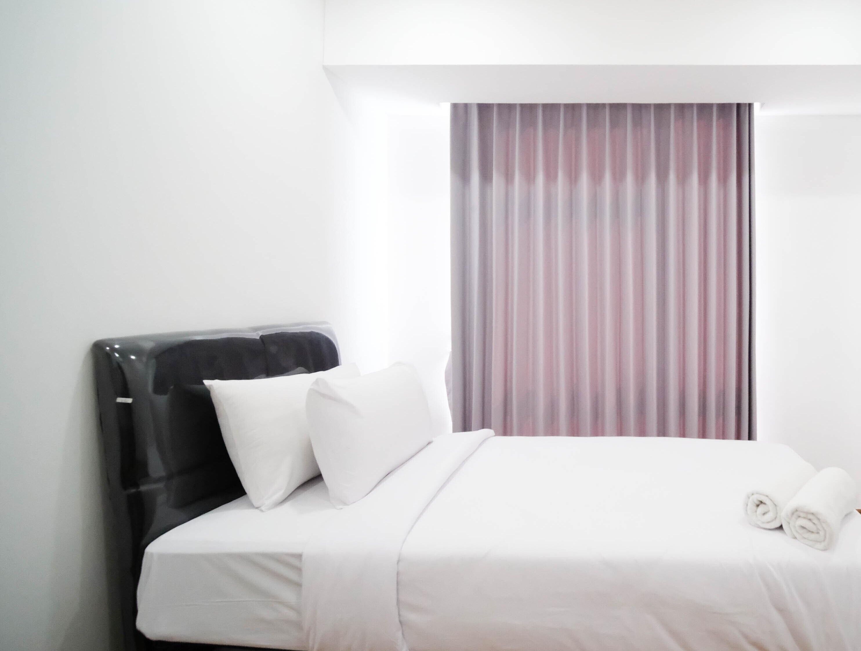 Una habitación de hotel con una cama, almohadas, un cabecero negro y un ventanal con cortinas.