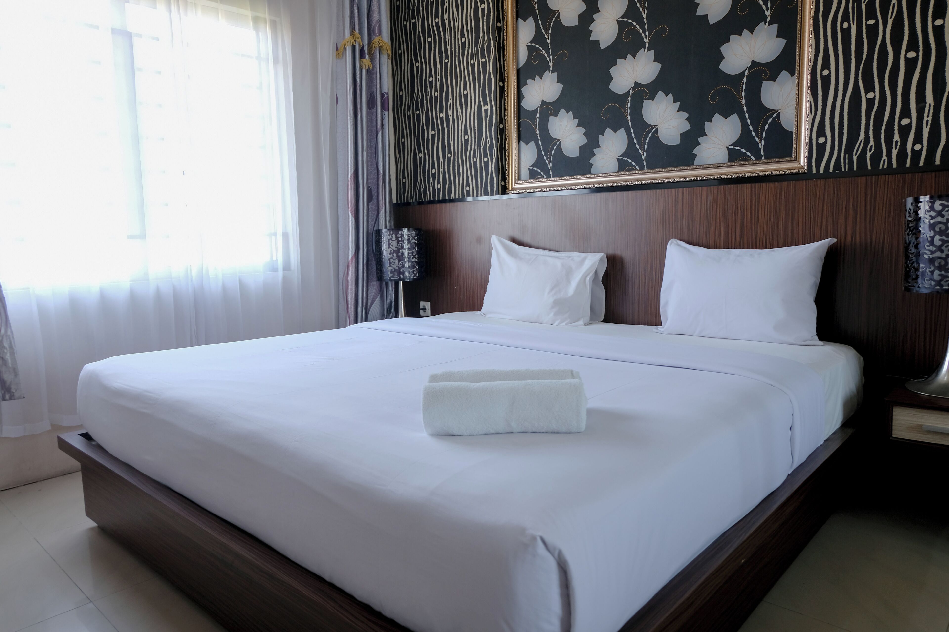 Una cama bien hecha con sábanas blancas y una toalla doblada, cabecera de madera y papel tapiz con estampado floral.