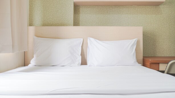A neatly made bed with white linens and two pillows, a wooden headboard, and a small bedside table with a lamp. - Cozy Stay 1Br At Green Pramuka City Apartment (Jakarta)