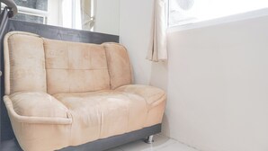 A beige sofa in a corner of a room with white walls and a window. - Cozy Stay 1Br At Green Pramuka City Apartment (Jakarta)