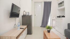 A compact living space with a TV mounted on the wall, a refrigerator, a wooden table with a bottle, a potted plant, and a grey sofa. - Nice And Tidy 2Br At Bassura City Apartment Near Mall (Jakarta)
