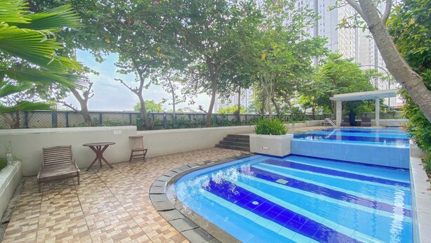 A swimming pool with a tiled deck, surrounded by greenery and a covered seating area. - Nice And Tidy 2Br At Bassura City Apartment Near Mall (Jakarta)
