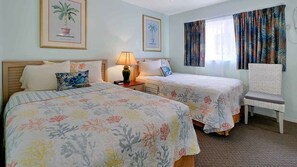 A hotel room with two beds, a chair, a nightstand, a lamp, and two framed pictures on the wall. - Carlton House Motel (St. Pete Beach)