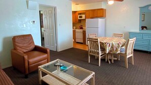 A living room with a brown leather chair, a glass coffee table, a dining table with chairs, a kitchen area with wooden cabinets, a white refrigerator, and a blue cabinet.