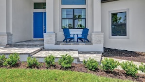 A modern house with a blue door, a covered patio with two blue chairs, and a small table. - Luxe Canal Home, Boat Rental, Walk to Cape Harbour (Cape Coral)