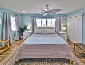 A bedroom with a bed, nightstands, a ceiling fan, a window with curtains, a plant, and a wooden floor.
