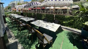 Un área de comedor al aire libre con mesas y sillas, rodeada de vegetación y edificios.