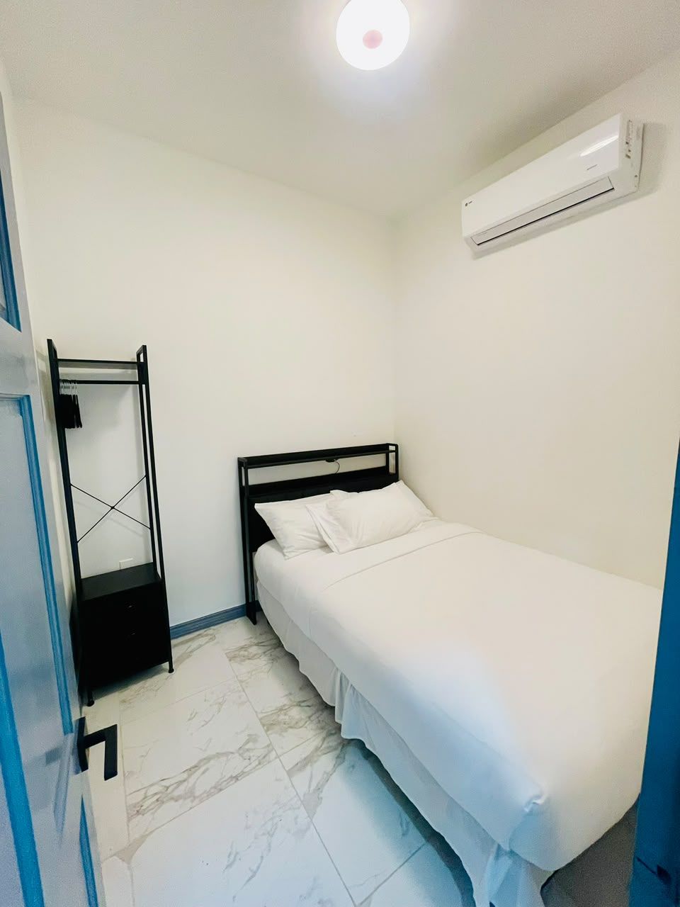 Een kleine eenpersoonskamer met een wit bed, een zwart metalen frame, een witte airconditioner aan de muur en een betegelde vloer.