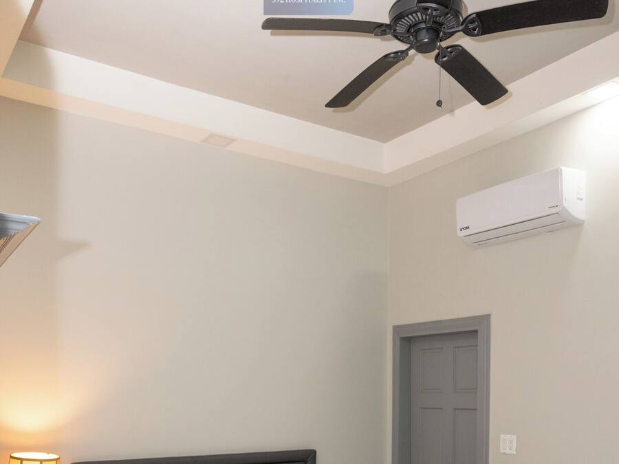 A neatly made bed with white linens, a bedside lamp, a ceiling fan, and an air conditioning unit on the wall.