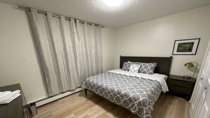 A bedroom with a bed, a nightstand, a lamp, a picture on the wall, and a curtain. - Stay Anchorage! Fully Furnished Apartments with GCI RED 2 GIG Internet (Anchorage)