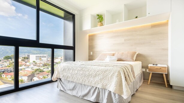 Ein modernes Schlafzimmer mit großem Fenster, einem Bett mit gemustertem Überwurf, einem Nachttisch und einem kleinen Beistelltisch.