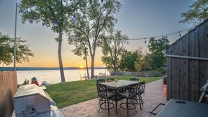 Une terrasse extérieure équipée d’un barbecue, d’une table à manger et de chaises, donnant sur un plan d’eau bordé d’arbres et délimité par une clôture en bois.