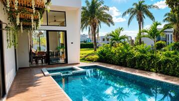 A modern outdoor pool area with a wooden deck, a glass door entrance, and lush greenery including palm trees.