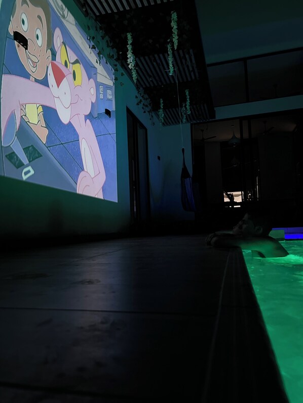 A room with a large animated character mural on the wall, a pool with green lighting, and a ceiling with hanging plants.