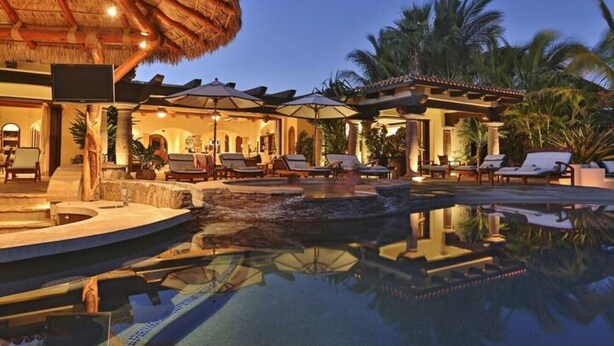 A luxurious pool area with lounge chairs and umbrellas, surrounded by palm trees and a thatched-roof structure. - Villa de Lujo en Cabo del Sol Casa Costa (Cabo San Lucas)