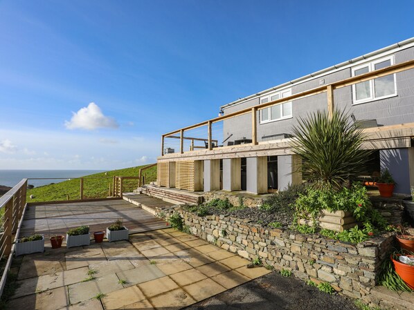A modern house with a stone wall and wooden deck, overlooking a grassy hill and the sea. - Bryn Y Mor (Tywyn)