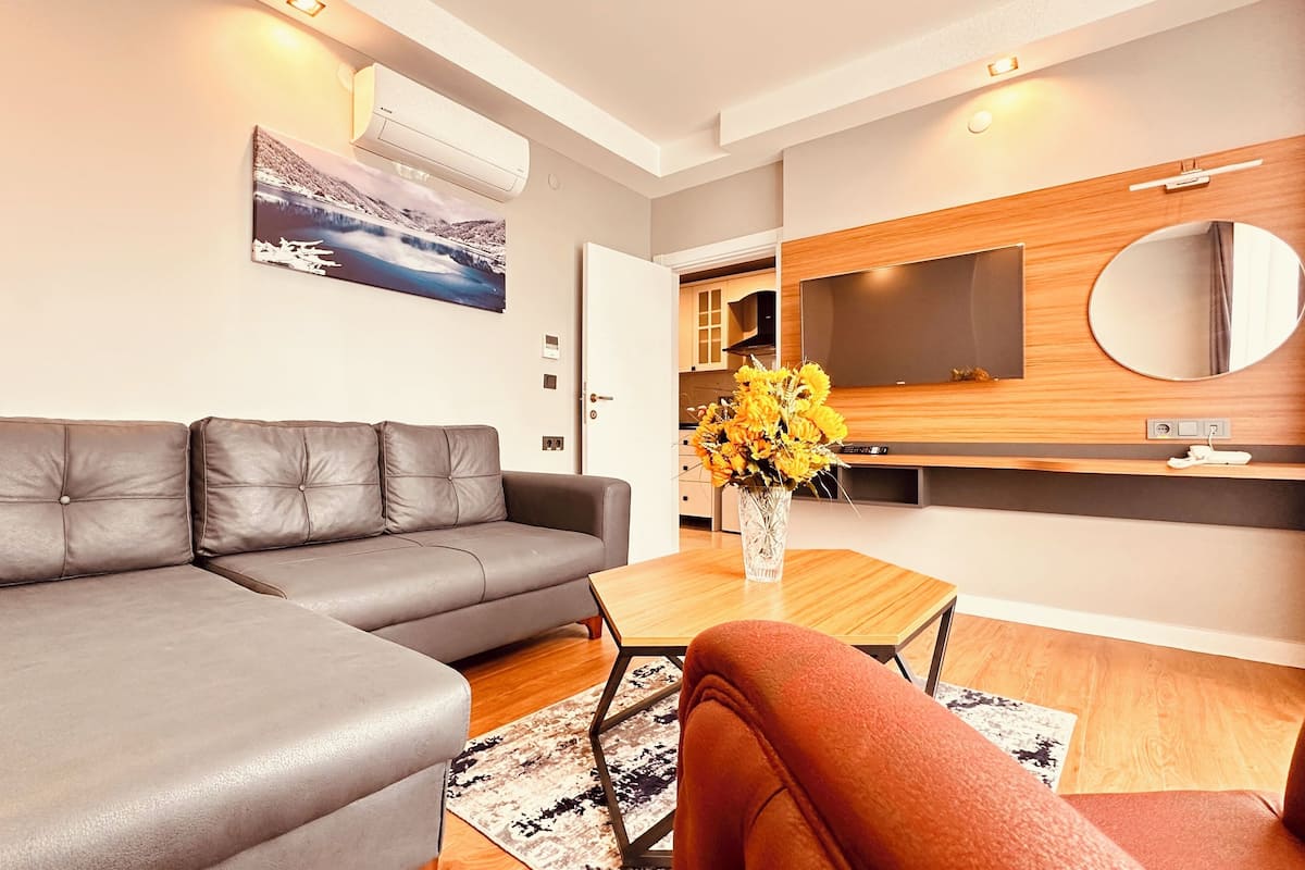 A modern living room with a grey sectional sofa, a wooden coffee table with a vase of flowers, and a large flat-screen TV mounted on a wooden wall.