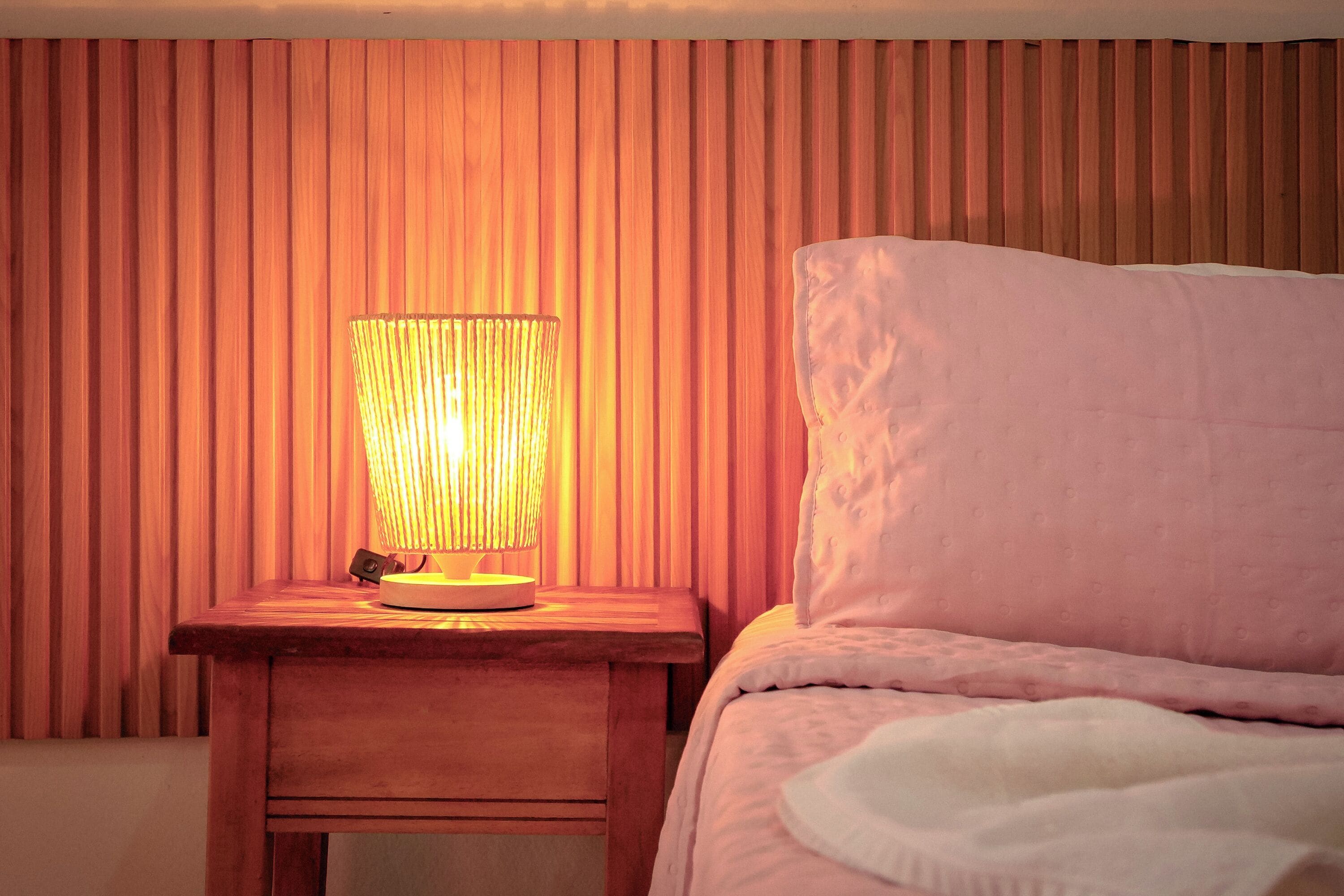 Una mesita de noche de madera con una lámpara, una cama con almohada blanca y una pared de madera con listones verticales.