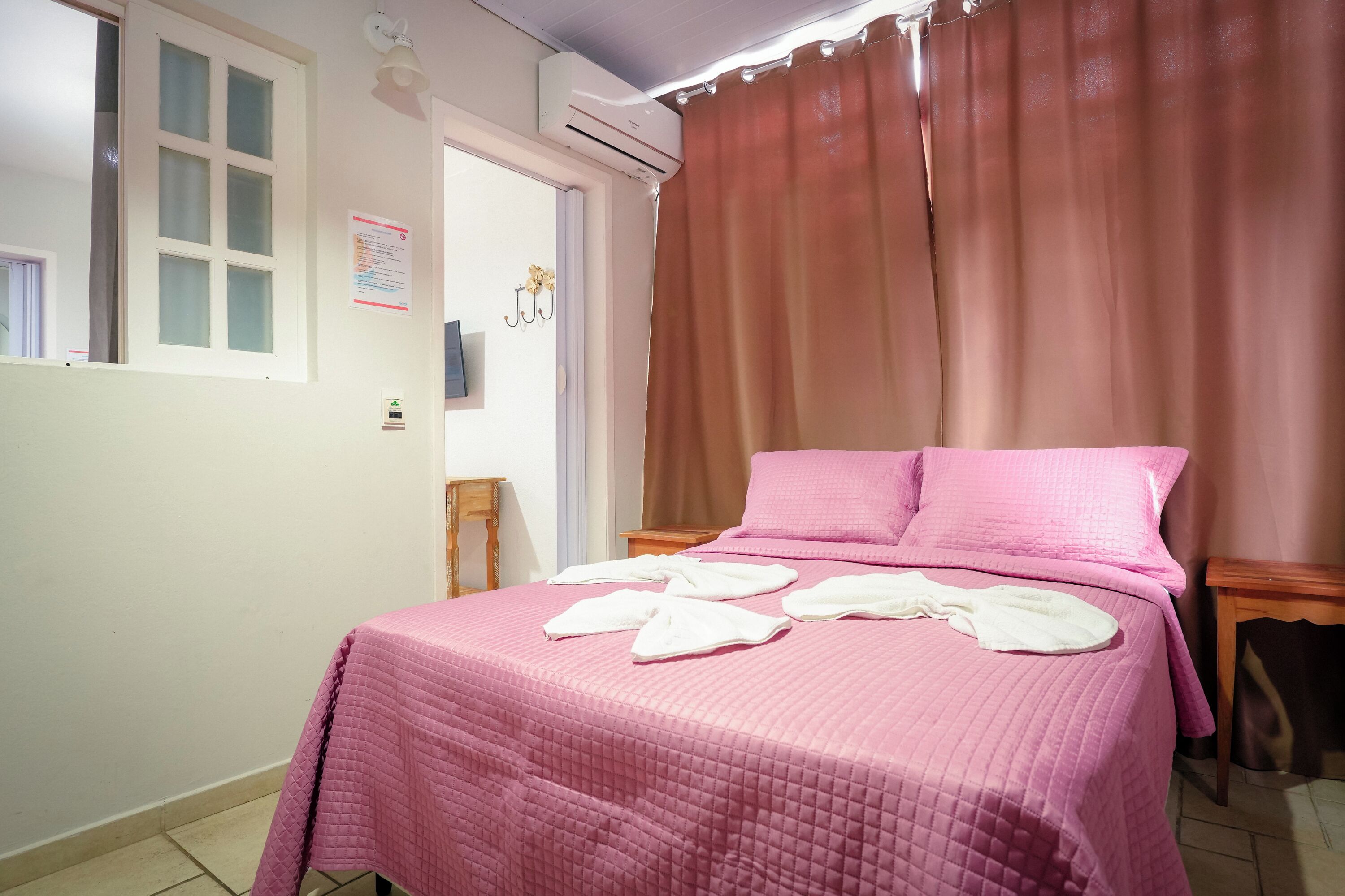 A bedroom with a pink quilted bedspread, a wooden nightstand, and a window with frosted glass.