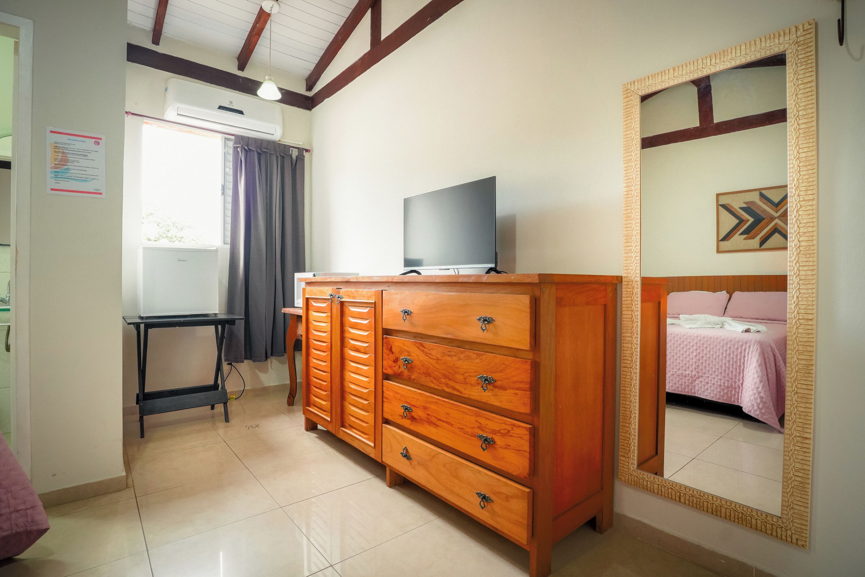 A room with a wooden dresser, a television on top, a mirror with a decorative frame, and a bed with pink bedding.