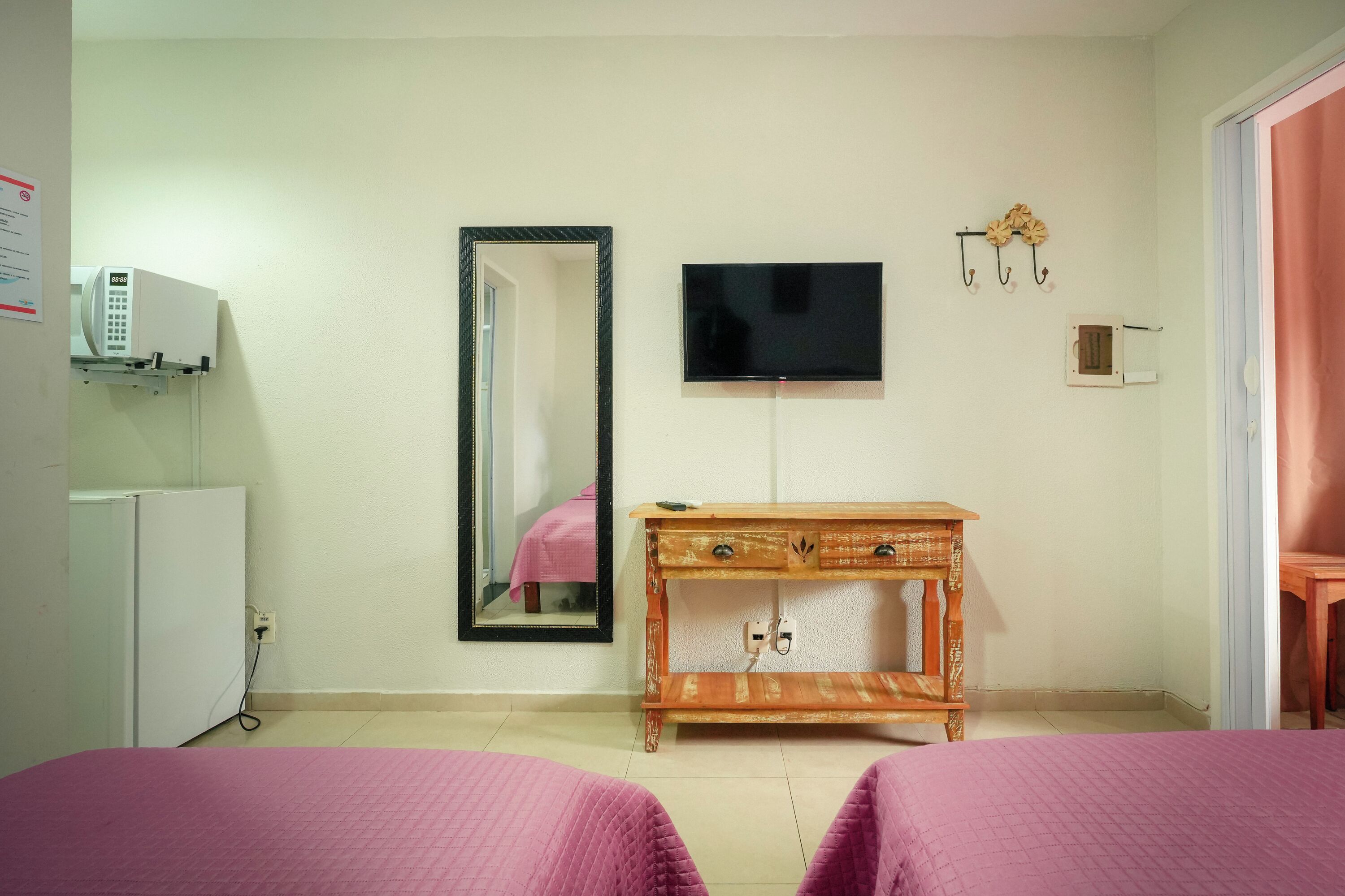 A bedroom with a bed, a TV, a mirror, a wooden dresser, and a white refrigerator.