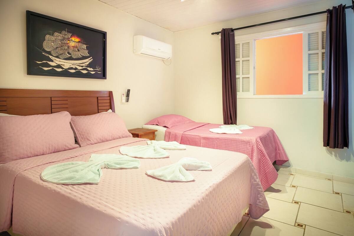 Une chambre d’hôtel avec un grand lit, deux petits lits, un tableau au mur, un climatiseur, une fenêtre avec des rideaux et une table de chevet avec une lampe.