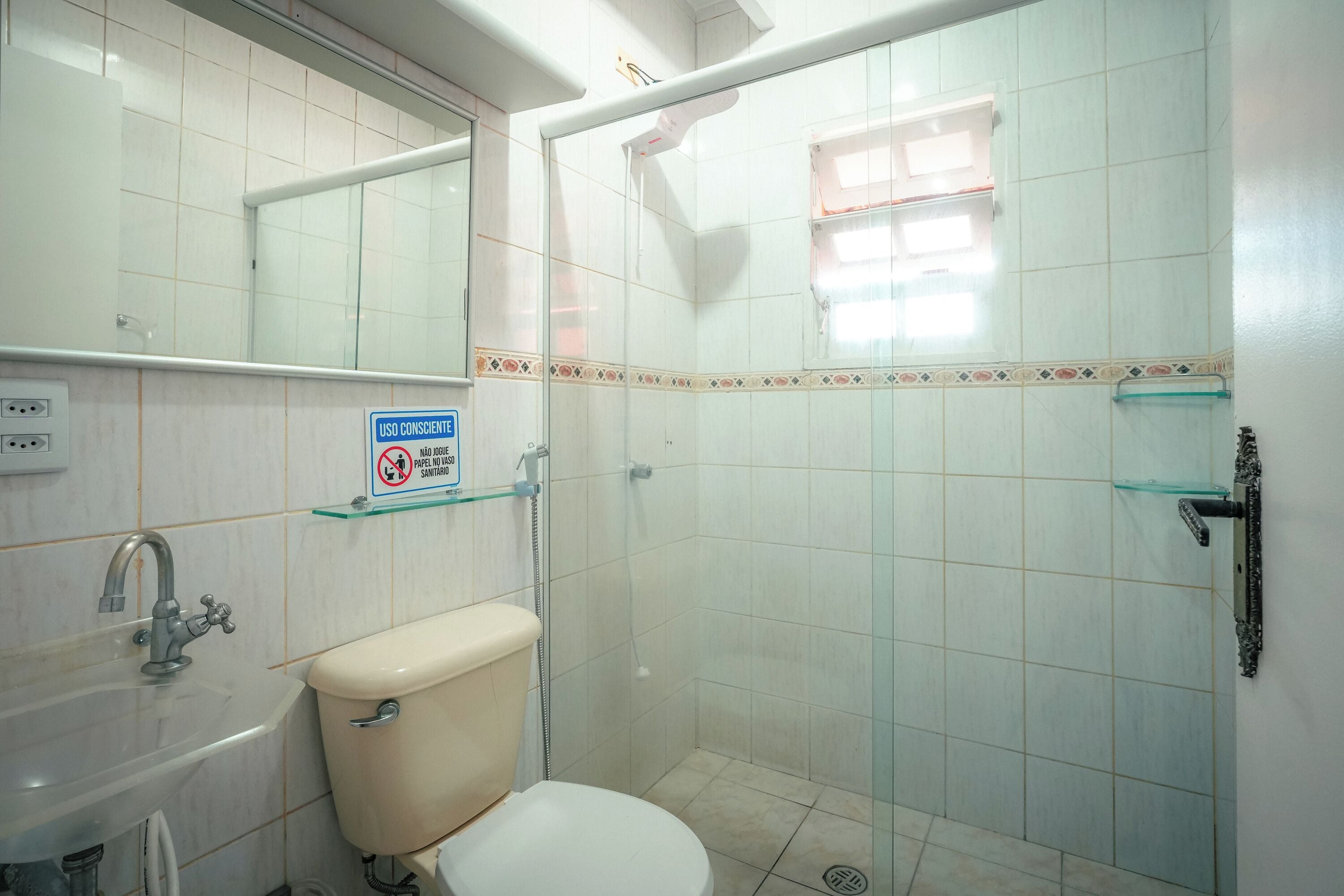 Baño con inodoro, lavabo y ducha. Hay un estante de vidrio sobre el lavabo y un espejo sobre el inodoro.