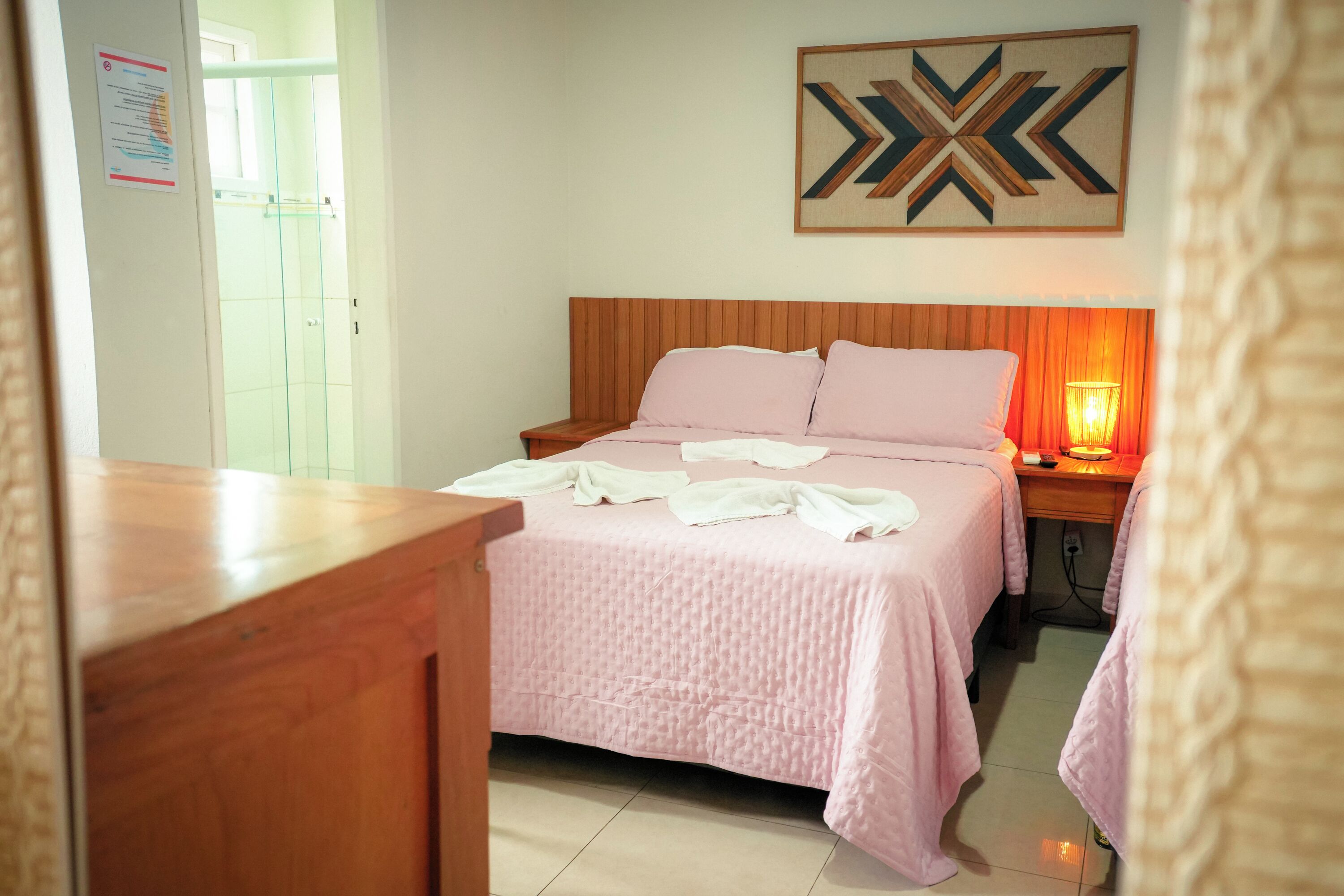 A hotel room with a wooden headboard, a bed with pink bedding, a bedside table with a lamp, and a wall-mounted abstract artwork.