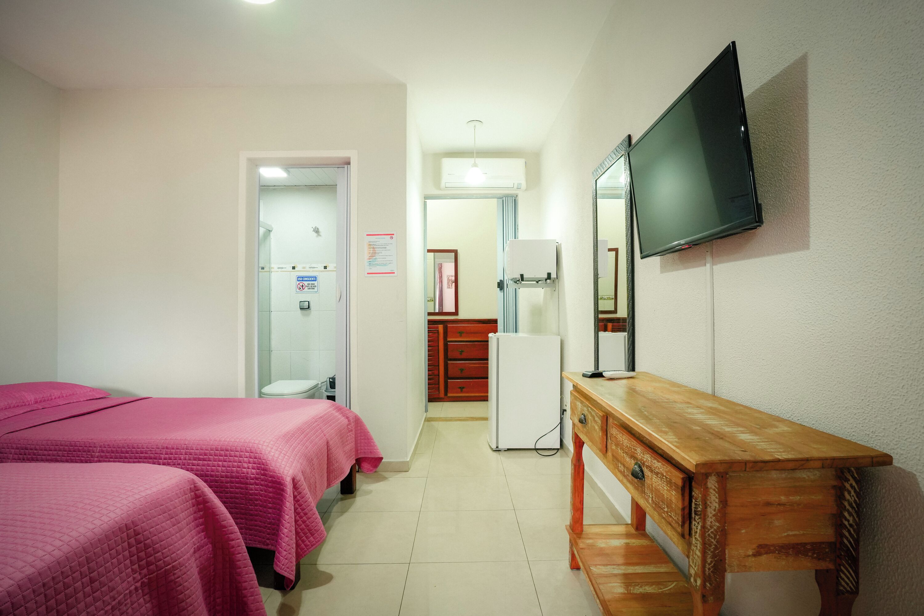 Habitación de hotel con dos camas, un aparador de madera, televisión, refrigerador y baño a la vista a través de una puerta abierta.