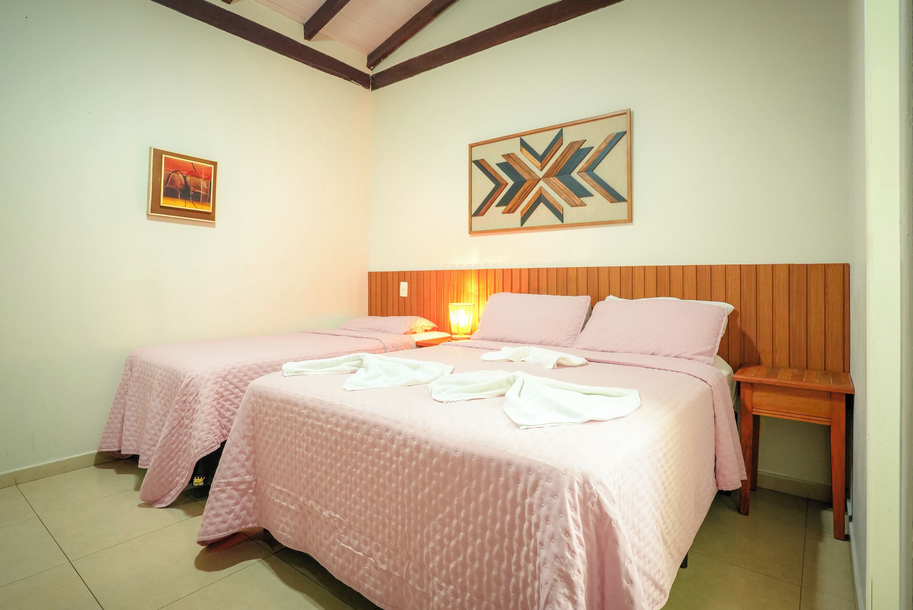 A hotel room with two beds, a wooden headboard, a wall-mounted artwork, and a bedside table with a lamp.