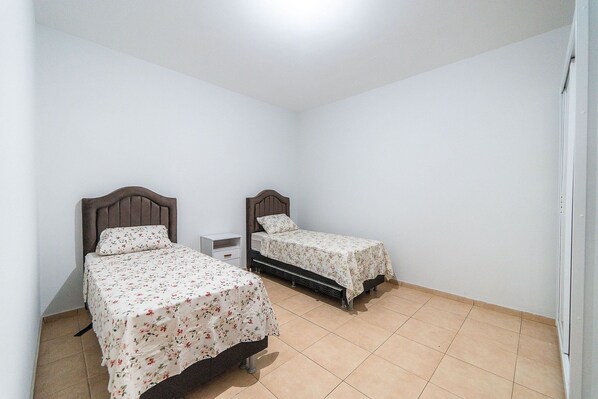 Una habitación con dos camas individuales, cada una con cabecera de madera y mesita de noche blanca. Las paredes son blancas y el piso está cubierto de baldosas.