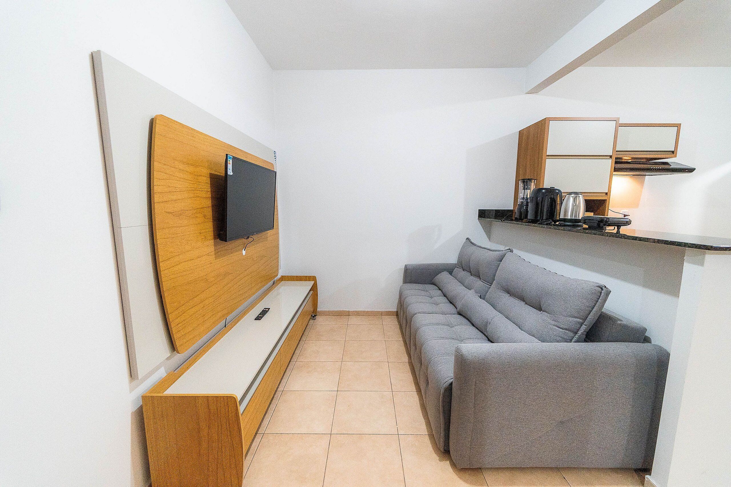 Un espacio habitable compacto con una TV de pantalla plana montada en un panel de madera, un sofá gris, una pequeña mesa de centro y una zona de kitchenette con microondas y hervidor.