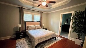 Ein Schlafzimmer mit einem Bett, Nachttischen, einem Deckenventilator, einem Fenster mit Jalousien und einer Pflanze.