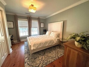 A bedroom with a bed, a nightstand, a lamp, a window with curtains, a ceiling light, and a plant.