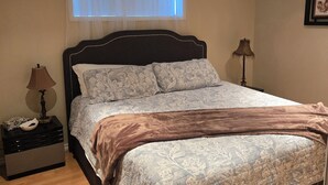 A bedroom with a bed, a nightstand, a lamp, a window with sheer curtains, and a framed picture on the wall.