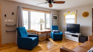A living room with a wooden floor, two blue armchairs, a wooden table, a TV on a white stand, and a ceiling fan. - Steps to Lake on the Mountain, 10Minutes to Picton (Prince Edward)