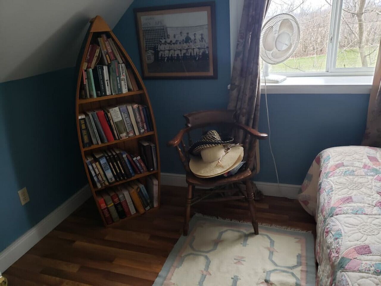 Une pièce avec une bibliothèque en bois, un fauteuil à bascule surmonté d’un chapeau, un tableau encadré accroché au mur et un lit recouvert d’une couvre-lit à motifs.