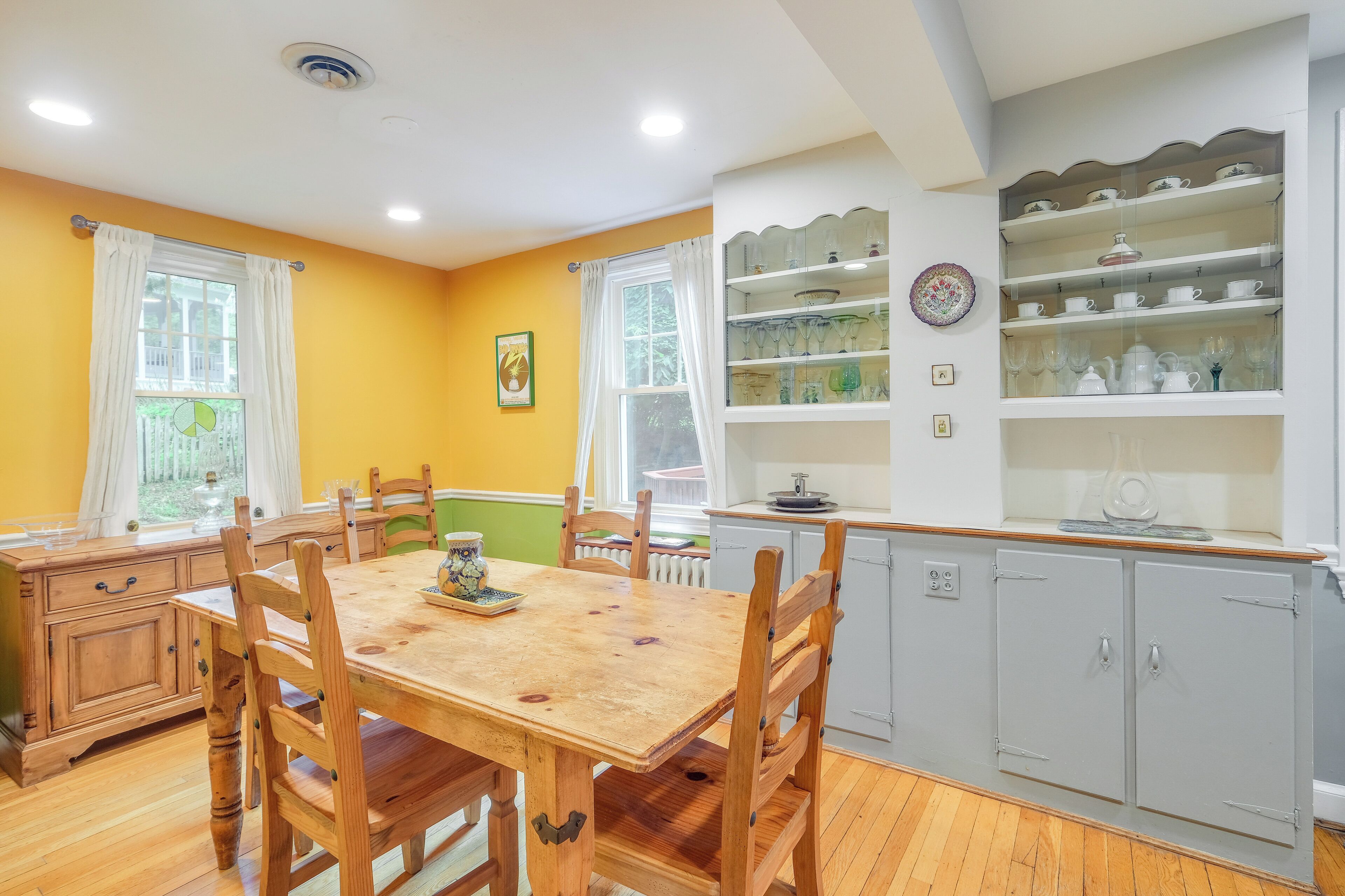 A dining area with a wooden table and chairs, a window with curtains, and a kitchen area with cabinets and shelves.