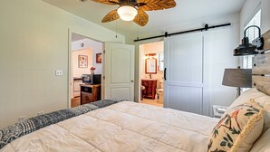 A bedroom with a bed, a ceiling fan, a bedside table, a lamp, and a view of the bathroom. - Fishing Pond & Pier: Secluded Broken Bow Cottage! (Broken Bow)