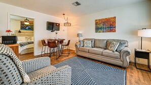 A living room with a sofa, armchair, dining table, and chairs. There is a TV mounted on the wall and a painting above the sofa. - Fishing Pond & Pier: Secluded Broken Bow Cottage! (Broken Bow)