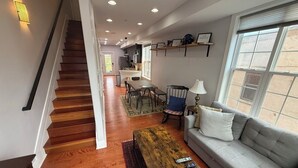 A spacious living area with a wooden staircase, a grey sofa, a coffee table, and a dining area with chairs and a table.