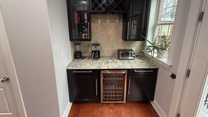 A compact kitchen with dark cabinets, a countertop with a coffee maker, toaster, and wine glasses, and a window with a plant.