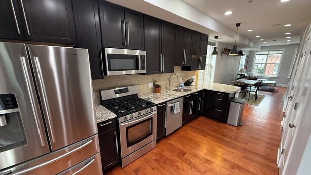 A modern kitchen with dark cabinets, stainless steel appliances, and a wooden floor.