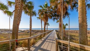 A wooden boardwalk with metal railings, surrounded by palm trees and leading to a beach.