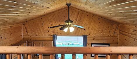 A wooden cabin interior with a balcony, a ceiling fan, and a view of the outdoors through a window.
