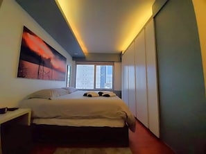 Ein modernes Schlafzimmer mit einem großen Bett, einem Nachttisch, einem Gemälde und einem Fenster mit Blick auf die Stadt.