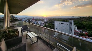 Een balkon met rieten meubels, een tafel en uitzicht op een stadsbeeld bij zonsondergang.