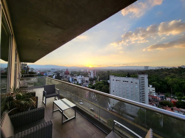 Ein Balkon mit Korbmöbeln, einem Tisch und Blick auf eine Stadtlandschaft bei Sonnenuntergang.