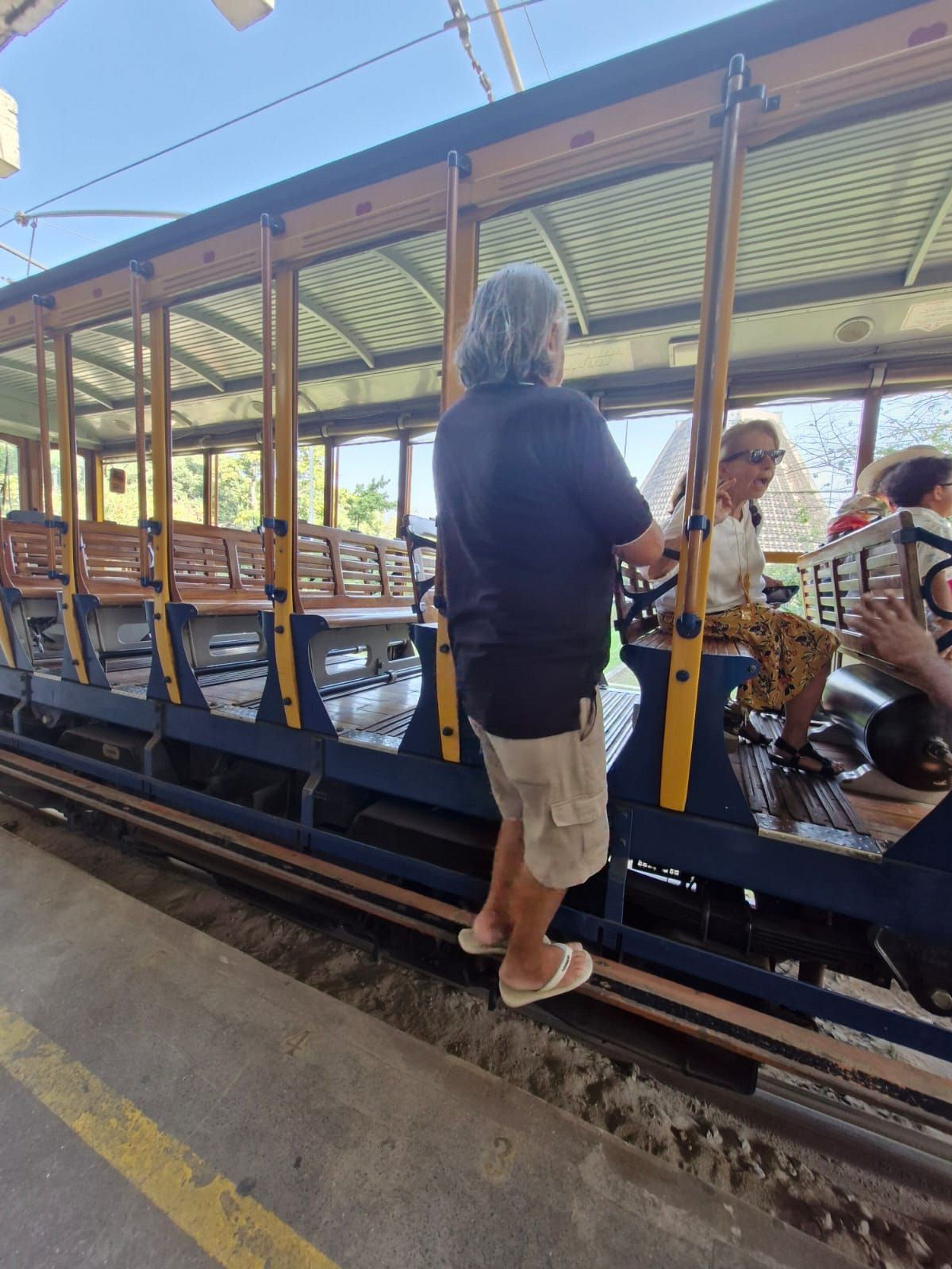 A vintage train with wooden benches and metal poles, passengers boarding at a station platform.